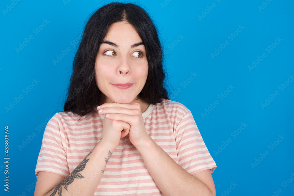 Curious young beautiful tattooed girl wearing pink striped t-shirt standing against blue background keeps hands under chin bites lips and looks with interest aside. Curious young beautiful tattooed girl wearing pink striped t-shirt standing against blue background keeps hands under chin bites lips and looks with interest aside.
