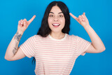 Cheerful young beautiful tattooed girl wearing pink striped t-shirt standing against blue background demonstrating hairdo