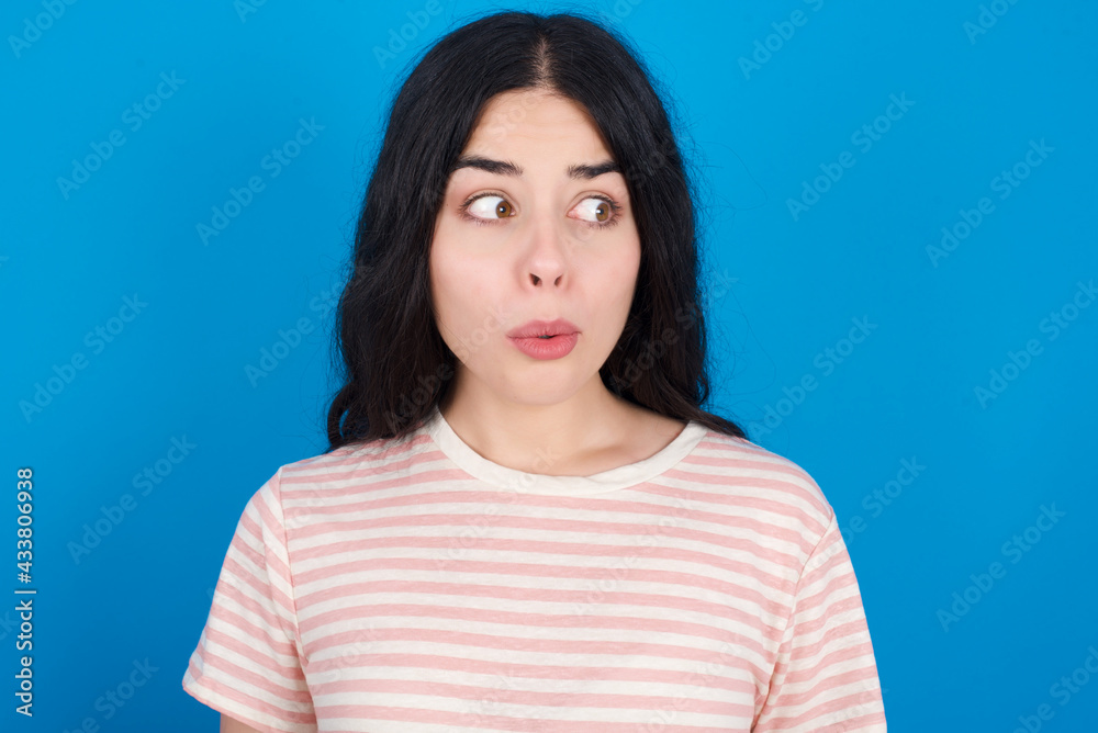 Shocked young beautiful tattooed girl wearing pink striped t-shirt standing against blue background look empty space with open mouth screaming: Oh My God! I can't believe this. Shocked young beautiful tattooed girl wearing pink striped t-shirt standing against blue background look empty space with open mouth screaming: Oh My God! I can't believe this.