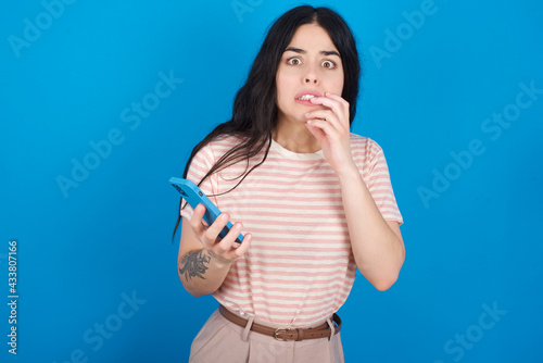 Afraid funny young beautiful tattooed girl wearing pink striped t-shirt standing against blue background holding telephone and bitting nails
