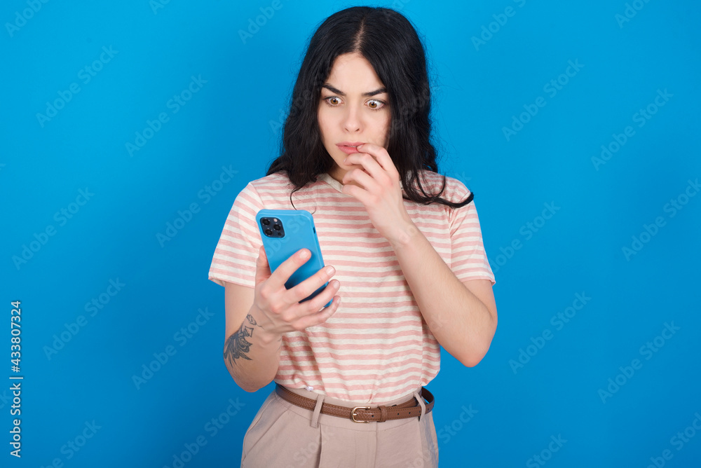 Portrait of pretty frightened young beautiful tattooed girl wearing pink striped t-shirt standing against blue background chatting biting nails after reading some scary news on her smartphone. Portrait of pretty frightened young beautiful tattooed girl wearing pink striped t-shirt standing against blue background chatting biting nails after reading some scary news on her smartphone.