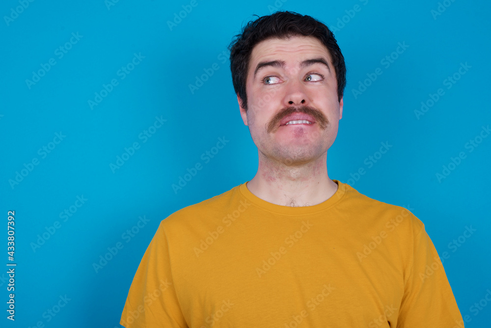 Coquettish young handsome Caucasian man with moustache wearing orange t-shirt against blue background smiling happily, blinking at camera in a playful manner, flirting with you. Coquettish young handsome Caucasian man with moustache wearing orange t-shirt against blue background smiling happily, blinking at camera in a playful manner, flirting with you.