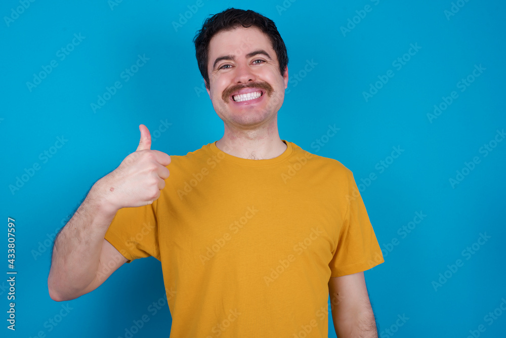 young handsome Caucasian man with moustache wearing orange t-shirt against blue background doing happy thumbs up gesture with hand. Approving expression looking at the camera showing success. young handsome Caucasian man with moustache wearing orange t-shirt against blue background doing happy thumbs up gesture with hand. Approving expression looking at the camera showing success.