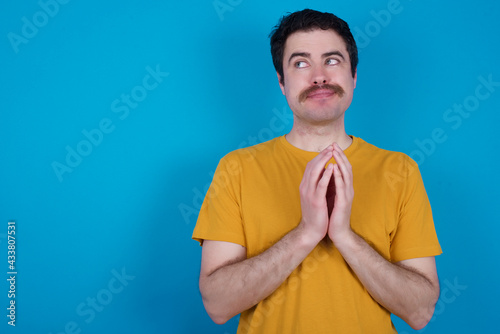 Charming cheerful young handsome Caucasian man with moustache wearing orange t-shirt against blue background making up plan in mind holding hands together, setting up an idea. Look askance