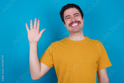 young handsome Caucasian man with moustache wearing orange t-shirt against blue background Waiving saying hello happy and smiling, friendly welcome gesture.