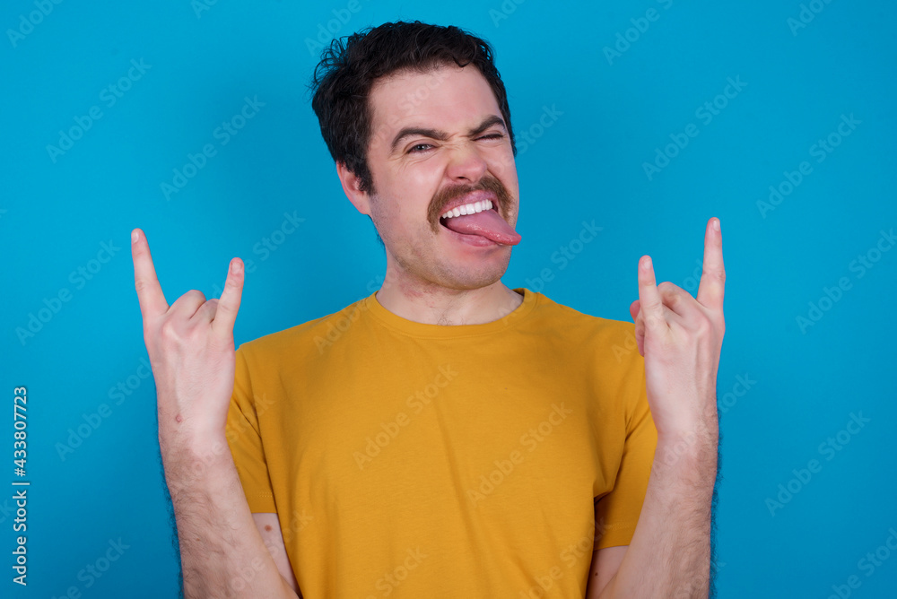 young handsome Caucasian man with moustache wearing orange t-shirt against blue background making rock hand gesture and showing tongue young handsome Caucasian man with moustache wearing orange t-shirt against blue background making rock hand gesture and showing tongue