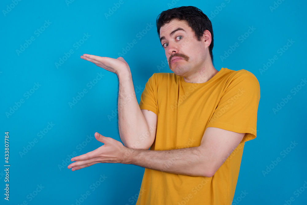 young handsome Caucasian man with moustache wearing orange t-shirt against blue pointing aside with both hands showing something strange and saying: I don't know what is this. Advertisement concept. young handsome Caucasian man with moustache wearing orange t-shirt against blue pointing aside with both hands showing something strange and saying: I don't know what is this. Advertisement concept.