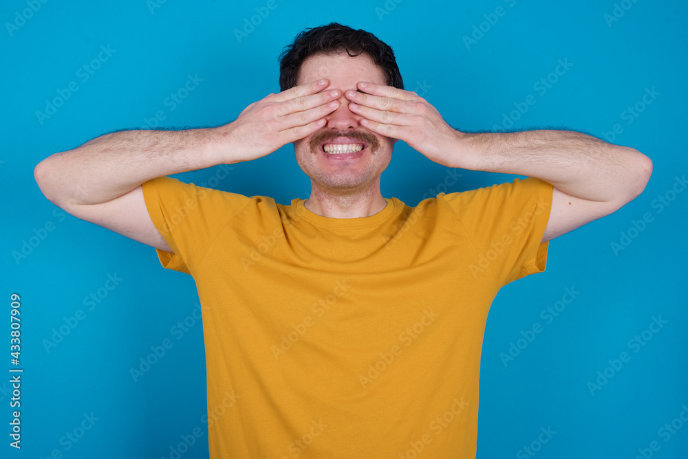 young handsome Caucasian man with moustache wearing orange t-shirt against blue background covering eyes with hands smiling cheerful and funny. Blind concept.