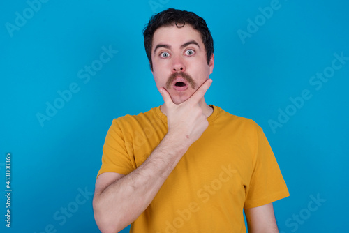 young handsome Caucasian man with moustache wearing yellow t-shirt against blue background Looking fascinated with disbelief, surprise and amazed expression with hands on chin