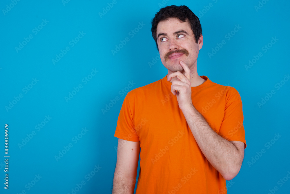 Thoughtful young handsome Caucasian man with moustache wearing orange t-shirt against blue background holds chin and looks away pensively makes up great plan