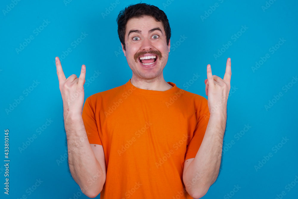 young handsome Caucasian man with moustache wearing orange t-shirt against blue background makes rock n roll sign looks self confident and cheerful enjoys cool music at party. Body language concept. young handsome Caucasian man with moustache wearing orange t-shirt against blue background makes rock n roll sign looks self confident and cheerful enjoys cool music at party. Body language concept.