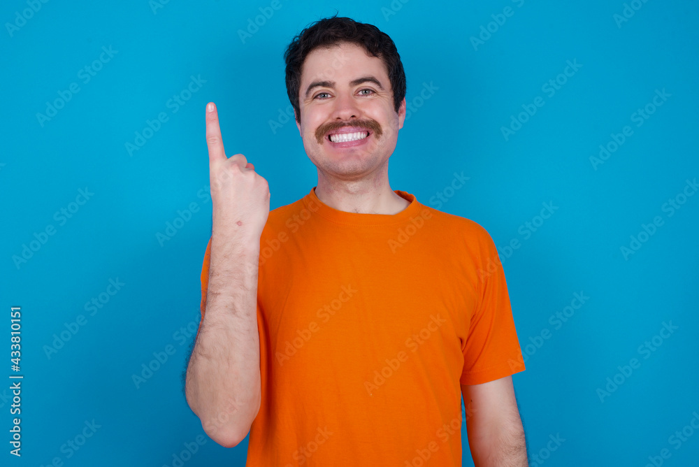 young handsome Caucasian man with moustache wearing orange t-shirt against blue background smiling and looking friendly, showing number one or first with hand forward, counting down young handsome Caucasian man with moustache wearing orange t-shirt against blue background smiling and looking friendly, showing number one or first with hand forward, counting down