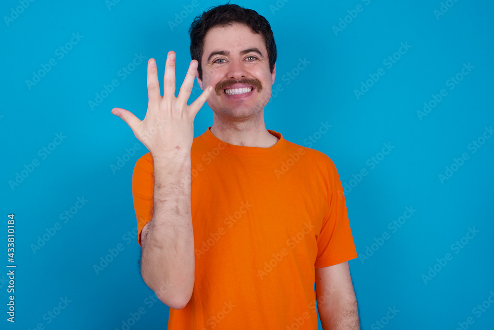 young handsome Caucasian man with moustache wearing orange t-shirt against blue background smiling and looking friendly, showing number five or fifth with hand forward, counting down young handsome Caucasian man with moustache wearing orange t-shirt against blue background smiling and looking friendly, showing number five or fifth with hand forward, counting down