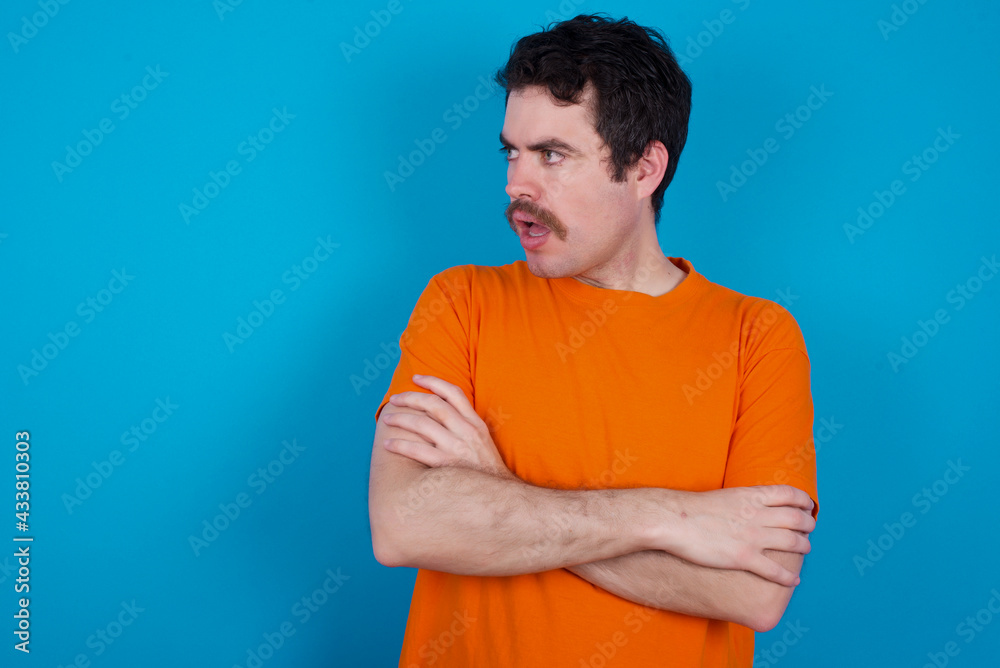 Astonished young handsome Caucasian man with moustache wearing orange t-shirt against blue background looks aside surprisingly with opened mouth. Astonished young handsome Caucasian man with moustache wearing orange t-shirt against blue background looks aside surprisingly with opened mouth.