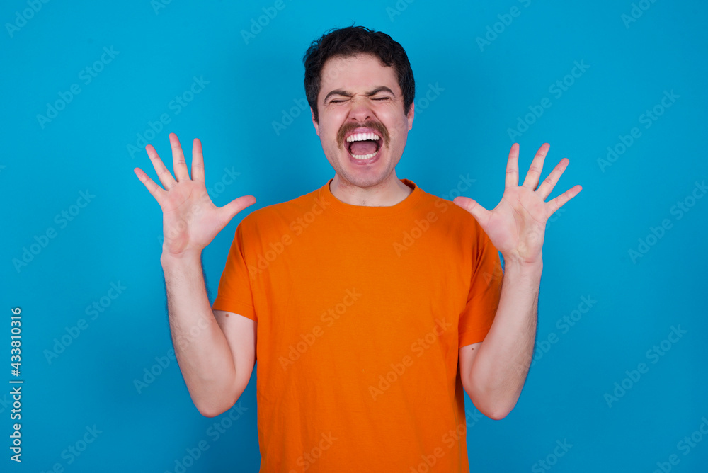 Crazy outraged young handsome Caucasian man with moustache wearing orange t-shirt against blue background screams loudly and gestures angrily yells furiously. Negative human emotions feelings concept Crazy outraged young handsome Caucasian man with moustache wearing orange t-shirt against blue background screams loudly and gestures angrily yells furiously. Negative human emotions feelings concept