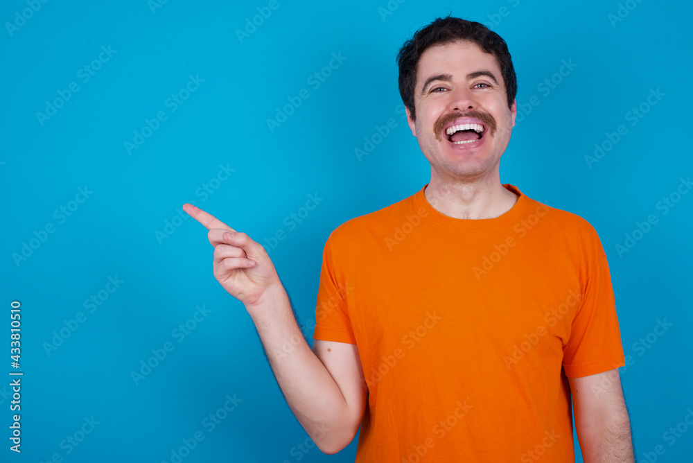 young handsome Caucasian man with moustache wearing orange t-shirt against blue laughs happily points away on blank space demonstrates shopping discount offer, excited by good news or unexpected sale. young handsome Caucasian man with moustache wearing orange t-shirt against blue laughs happily points away on blank space demonstrates shopping discount offer, excited by good news or unexpected sale.