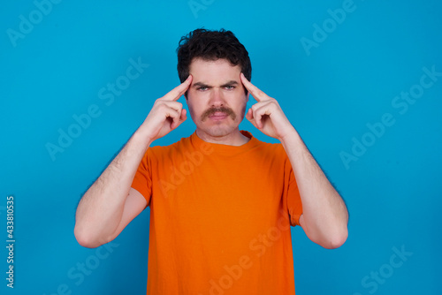 Serious concentrated young handsome Caucasian man with moustache wearing orange shirt keeps fingers on temples, tries to ease tension, gather with thoughts and remember important information for exam