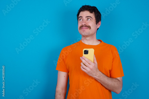 young handsome Caucasian man with moustache wearing orange t-shirt against blue holds mobile phone uses high speed internet and social networks has online communication. Modern technologies concept