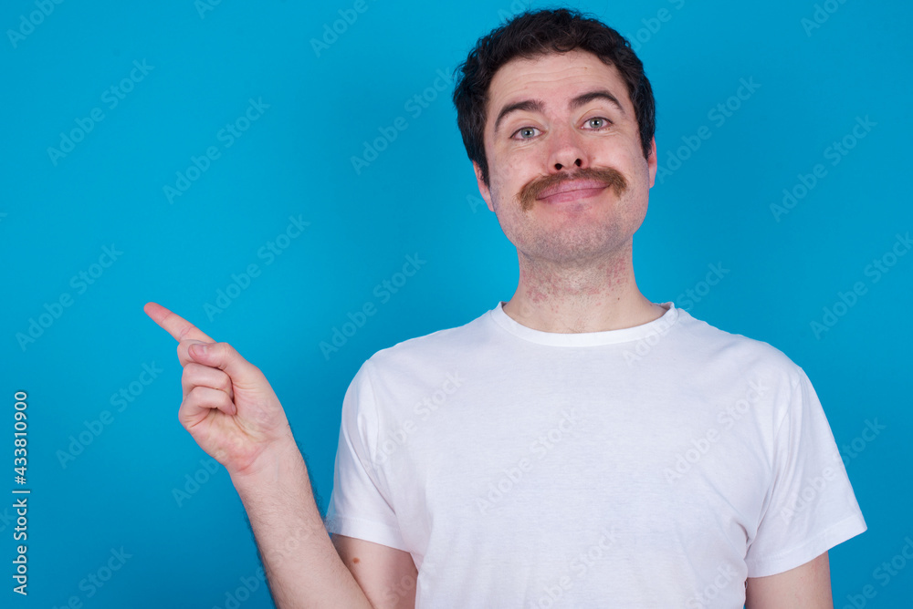 Positive young handsome Caucasian man with moustache wearing white t-shirt against blue background with satisfied expression indicates at upper right corner shows good offer suggests to click on link Positive young handsome Caucasian man with moustache wearing white t-shirt against blue background with satisfied expression indicates at upper right corner shows good offer suggests to click on link