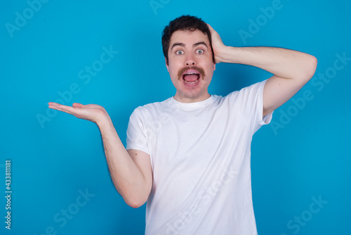 Shocked amazed surprised young handsome Caucasian man with moustache wearing white t-shirt against white background hold hand offering proposition