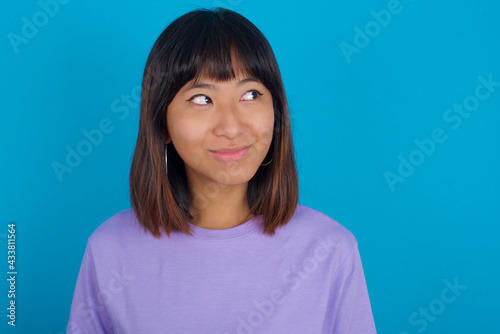 Young beautiful asian girl wearing purple t-shirt against blue background looking aside into empty space thoughtful