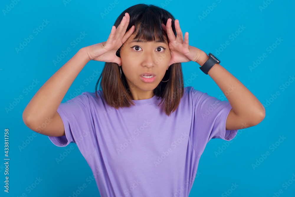 Young beautiful asian girl wearing purple t-shirt against blue background with scared expression, keeps hands on head, jaw dropped, has terrific expression. Omg concept Young beautiful asian girl wearing purple t-shirt against blue background with scared expression, keeps hands on head, jaw dropped, has terrific expression. Omg concept