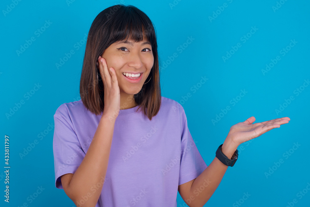 Crazy Young beautiful asian girl wearing purple t-shirt against blue background advising discount prices hold open palm new product Crazy Young beautiful asian girl wearing purple t-shirt against blue background advising discount prices hold open palm new product