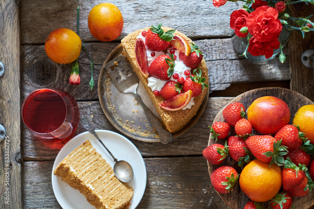 A piece of honey puff cake garnished with strawberries, red orange and cream. Flowers, wooden background, top view. A piece of honey puff cake garnished with strawberries, red orange and cream. Flowers, wooden background, top view.