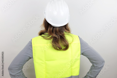 Female architect or engineer wearing reflecting jacket and hardhat standing backwards with hands on hips looking up.