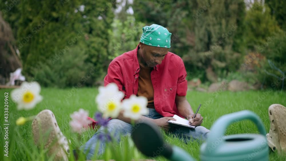 Front view of concentrated African American handsome man writing with pen on paper sitting on green lawn with blooming white flowers at front and summer garden at background. Landscaper planning work Front view of concentrated African American handsome man writing with pen on paper sitting on green lawn with blooming white flowers at front and summer garden at background. Landscaper planning work