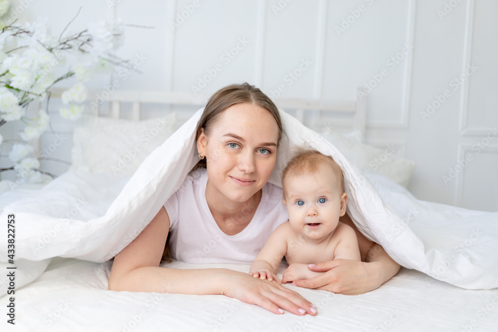 happy family, mom with a smiling baby under a blanket on a white cotton bed at home hugging happy family, mom with a smiling baby under a blanket on a white cotton bed at home hugging