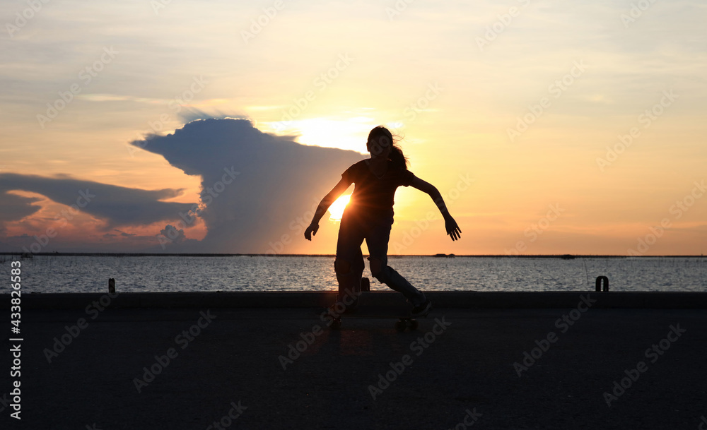 Young woman play surfskate at bridge along the seashore against sun set over the sea at Chonburi, Thailand