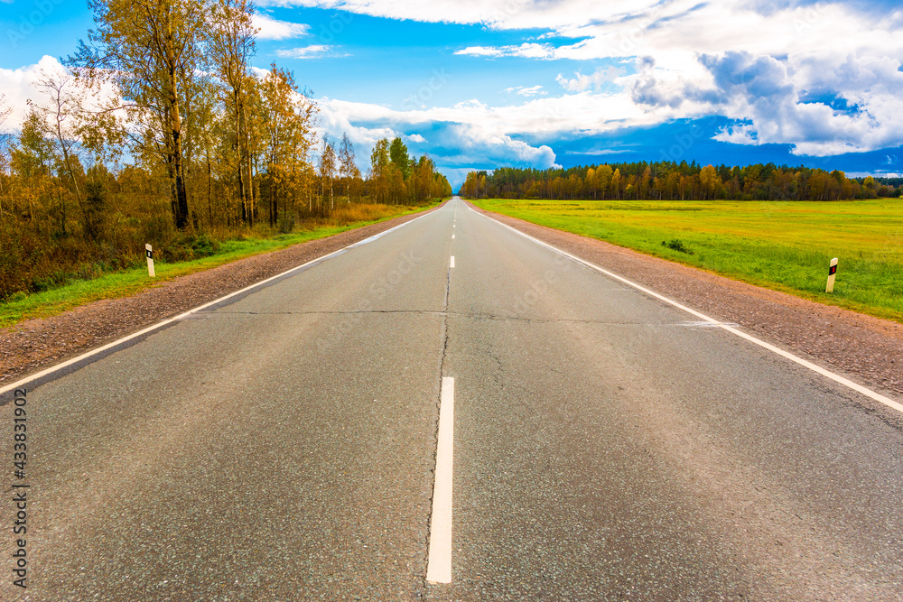 Fototapeta premium Autumn sky in the clouds lit by the sun over the empty road and boundless fields of the forest. Wide angle view