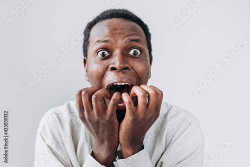 Portrait of a terrified African man staring in horror at the camera and putting his hands to his mouth in a light linen shirt on a white background. Very strong surprise or fright, horror in the eyes. photo