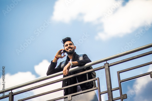 Happy bearded man standing outdoors while holding backpack and talking by smartphone