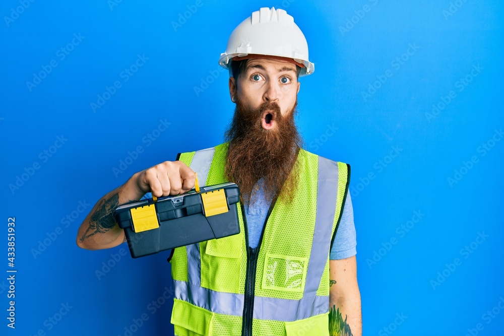 Redhead man with long beard wearing safety helmet and reflective jacket holding toolbox scared and amazed with open mouth for surprise, disbelief face Redhead man with long beard wearing safety helmet and reflective jacket holding toolbox scared and amazed with open mouth for surprise, disbelief face