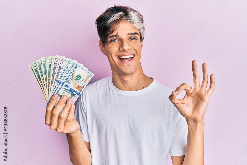 Young hispanic man holding dollars doing ok sign with fingers, smiling friendly gesturing excellent symbol Young hispanic man holding dollars doing ok sign with fingers, smiling friendly gesturing excellent symbol
