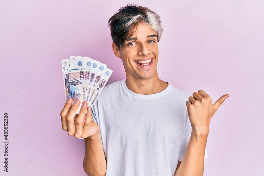 Young hispanic man holding russian 500 ruble banknotes pointing thumb up to the side smiling happy with open mouth Young hispanic man holding russian 500 ruble banknotes pointing thumb up to the side smiling happy with open mouth