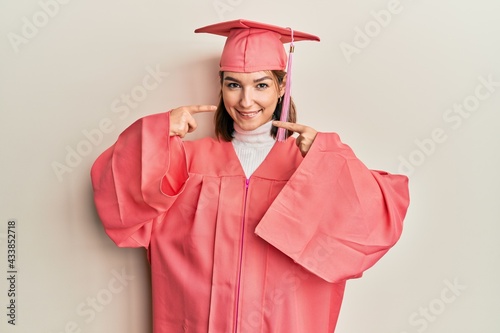 Young caucasian woman wearing graduation cap and ceremony robe smiling cheerful showing and pointing with fingers teeth and mouth. dental health concept.