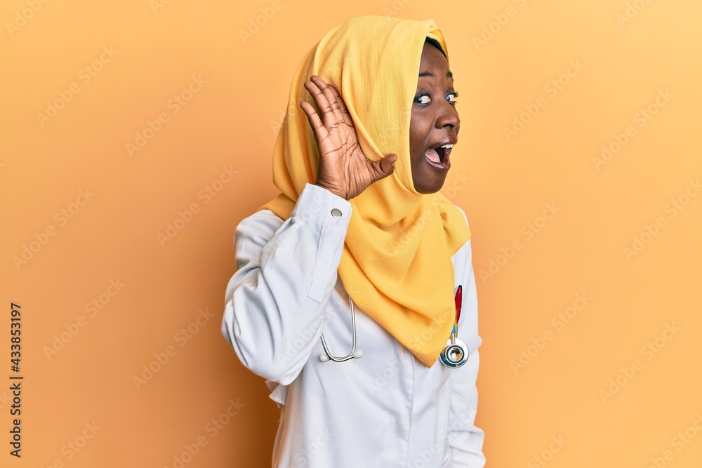 Beautiful african young woman wearing doctor uniform and hijab smiling with hand over ear listening an hearing to rumor or gossip. deafness concept. Beautiful african young woman wearing doctor uniform and hijab smiling with hand over ear listening an hearing to rumor or gossip. deafness concept.