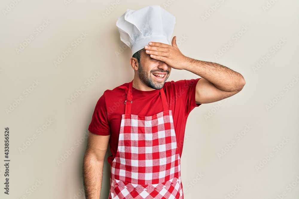 Young hispanic man wearing baker uniform smiling and laughing with hand on face covering eyes for surprise. blind concept. Young hispanic man wearing baker uniform smiling and laughing with hand on face covering eyes for surprise. blind concept.