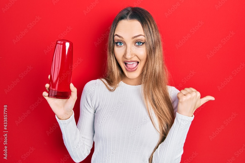 Naklejka premium Young blonde girl holding shampoo bottle pointing thumb up to the side smiling happy with open mouth