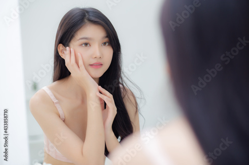 Beautiful young asian woman smiling look at mirror of checking face with skin care and cosmetic for rejuvenation and hygiene, beauty girl happy clean facial with cream or lotion and for health.