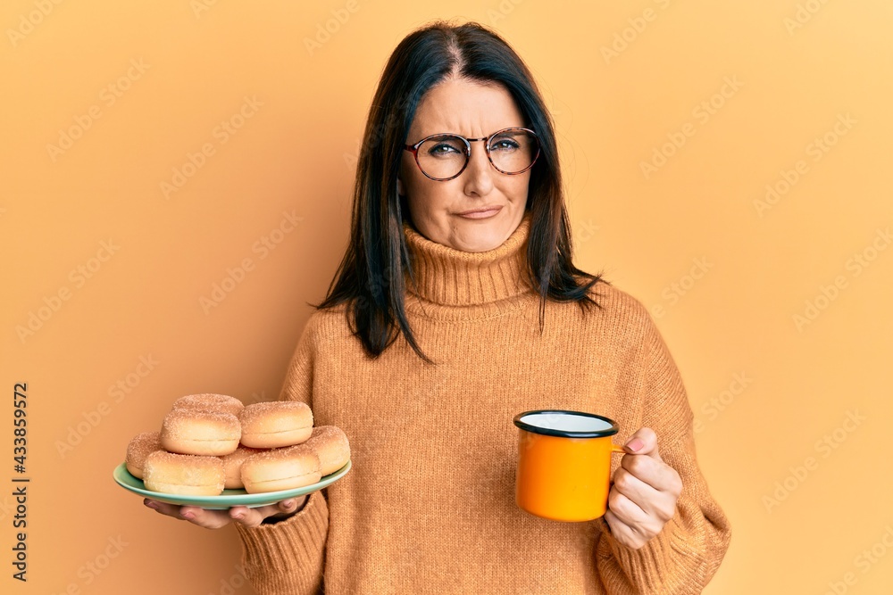 Middle age brunette woman eating doughnuts and drinking a cup of coffee skeptic and nervous, frowning upset because of problem. negative person. Middle age brunette woman eating doughnuts and drinking a cup of coffee skeptic and nervous, frowning upset because of problem. negative person.