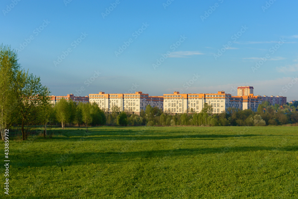 Panorama a park scene with a green grass field, a tree plant and a multi-storey residential complex against a blue sky