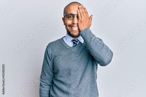 Hispanic adult man wearing glasses and business style covering one eye with hand, confident smile on face and surprise emotion. © Krakenimages.com