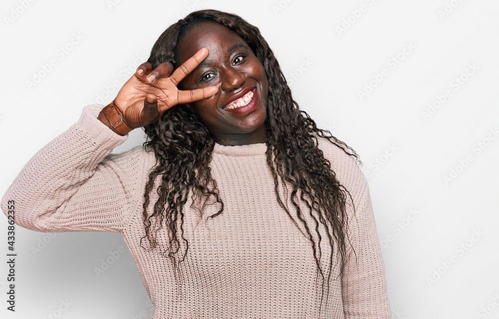 Young african woman wearing wool winter sweater doing peace symbol with fingers over face, smiling cheerful showing victory Young african woman wearing wool winter sweater doing peace symbol with fingers over face, smiling cheerful showing victory