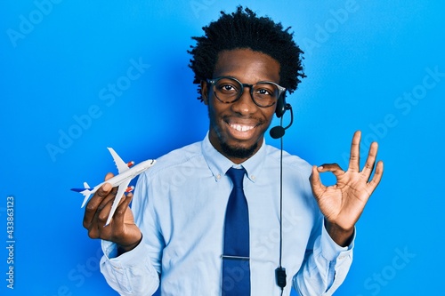 Young african american travel agent man holding plane doing ok sign with fingers, smiling friendly gesturing excellent symbol photo