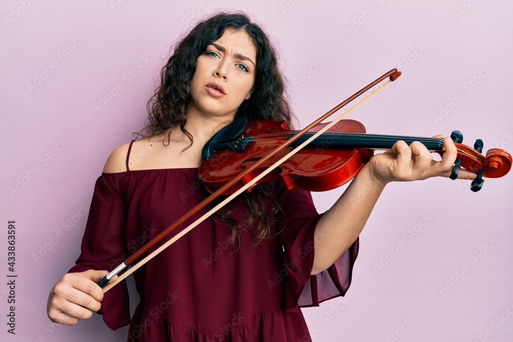 Young brunette musician woman with curly hair playing violin in shock face, looking skeptical and sarcastic, surprised with open mouth Young brunette musician woman with curly hair playing violin in shock face, looking skeptical and sarcastic, surprised with open mouth