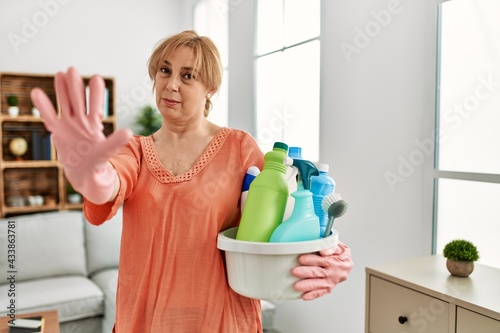 Middle age blonde woman holding cleaning products cleaning at home with open hand doing stop sign with serious and confident expression, defense gesture